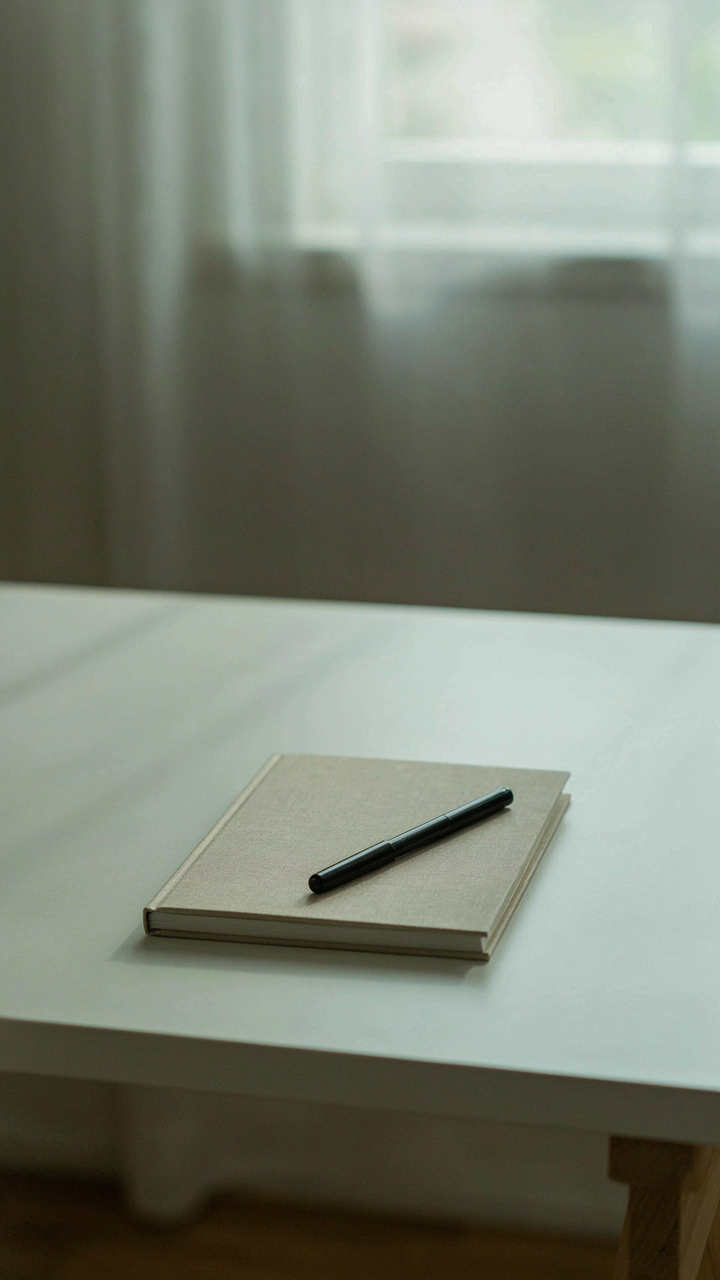 An empty white desk with a single notebook and pen in a sunny room.