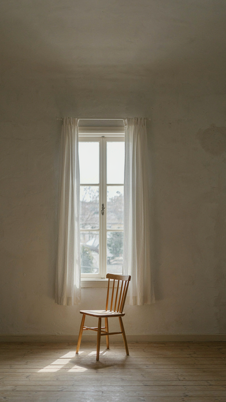 A minimalist bedroom wall with a window, sheer white curtains, and a wooden chair.