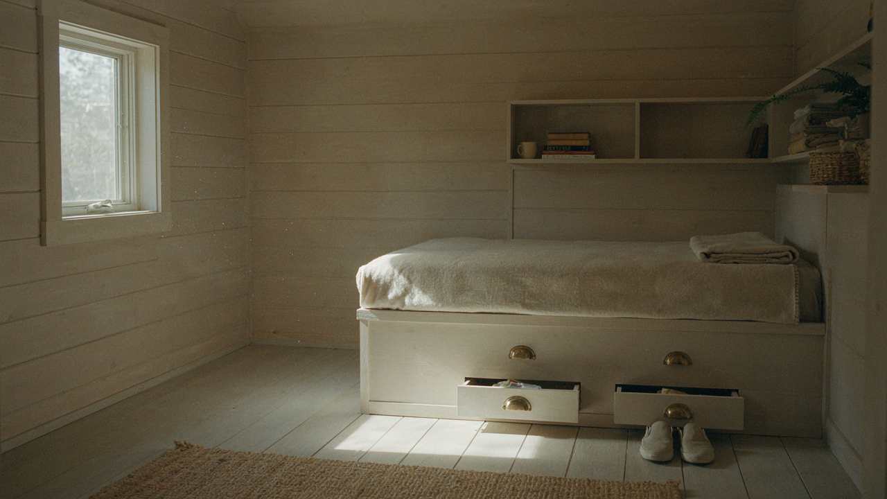 A tiny cabin bedroom with a built-in bed, integrated shelves, and warm sunlight.