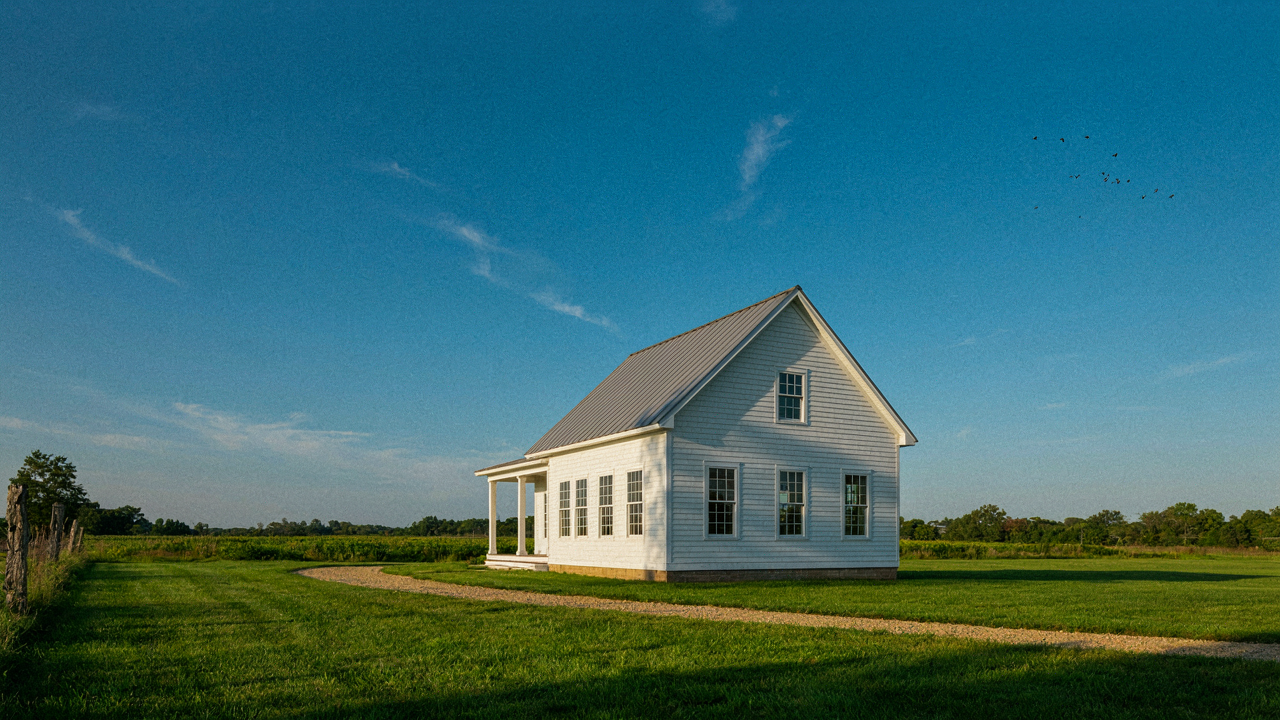 A classic white farmhouse with a metal roof stands in a green field.