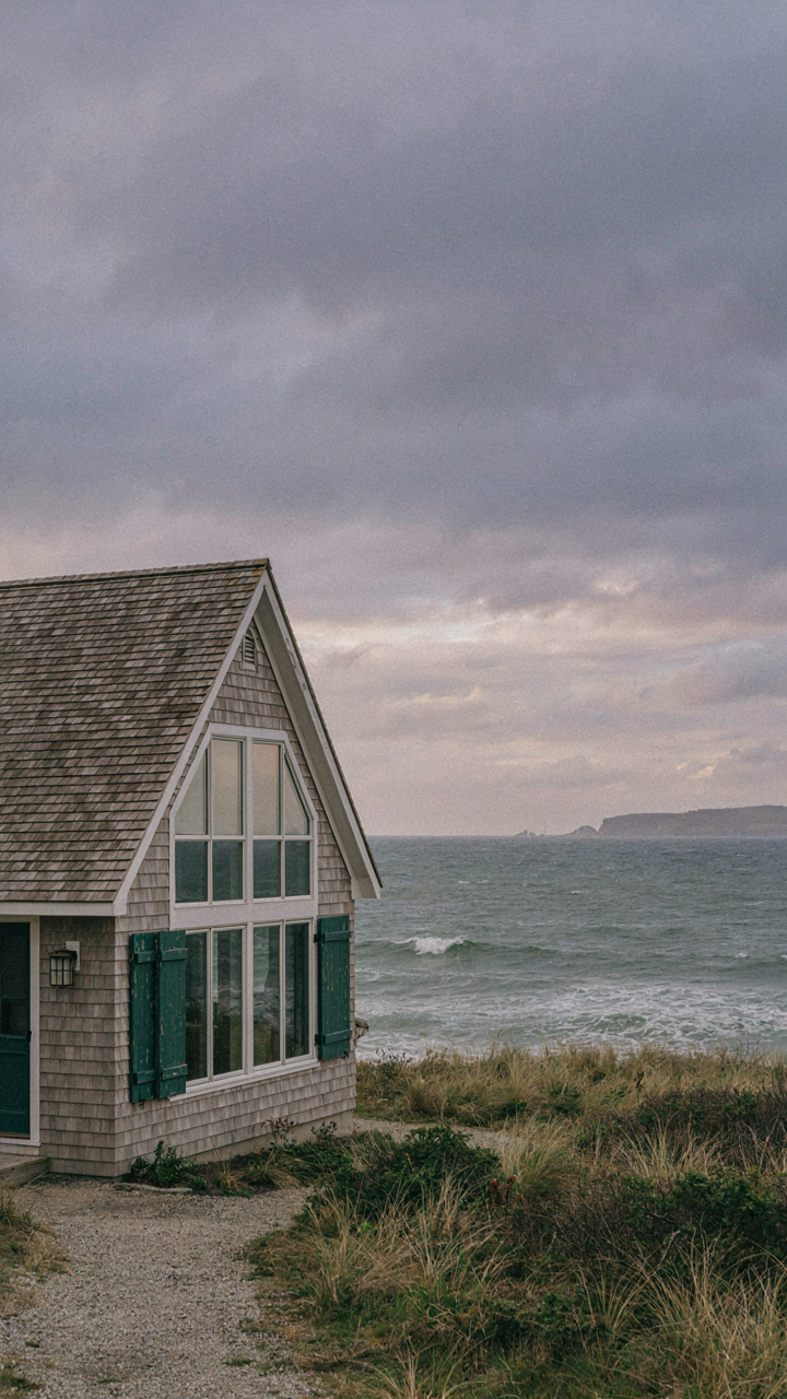 A modern cape cod style house has large picture windows facing a stormy ocean.