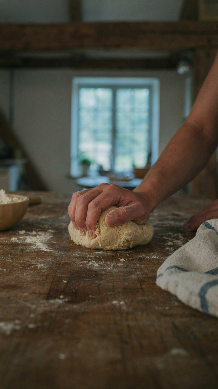 Hands knead dough on a worn butcher block counter in a kitchen.